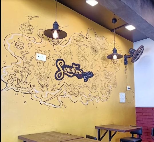 The marigold mural wall at Soushee Bites, covered in single-line drawings of fries, momos, soft serve and the shop's logo, under pendant lights.